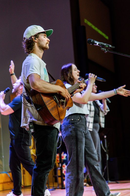 A band on stage: a man in a cap plays acoustic guitar beside singers with microphones