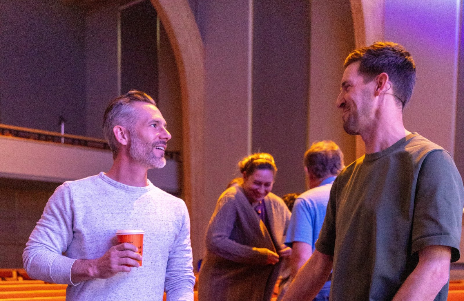 Two men talking and smiling in the sanctuary after a gathering, one holding a paper coffee cup, others in conversation behind them