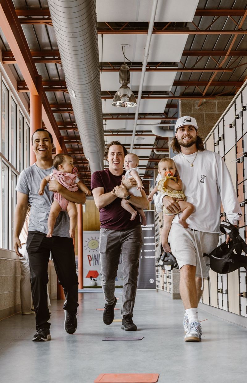 Three young dads walking down a hallway and laughing, each carrying a baby