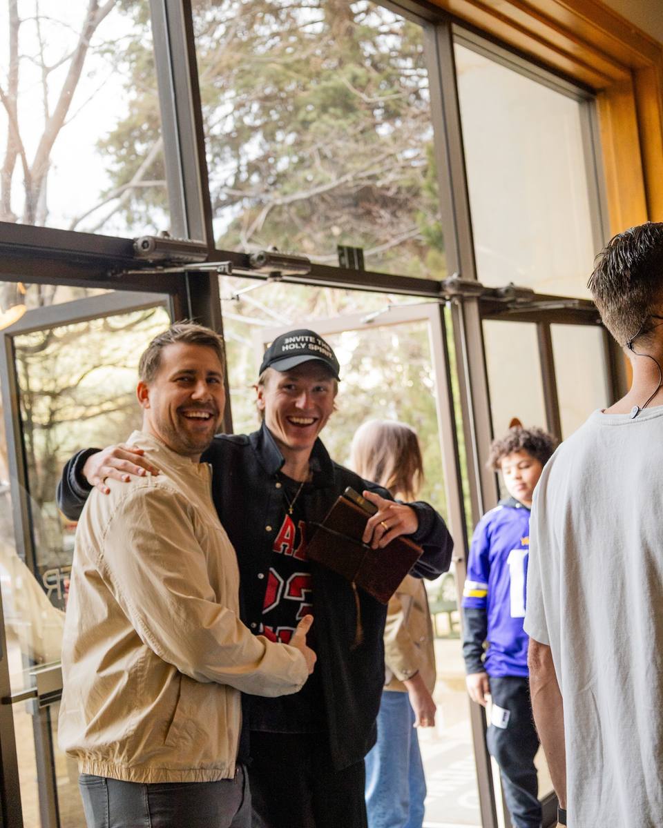 Two friends laughing with an arm around each other by the sunlit lobby doors, one in a cap holding a leather-bound book