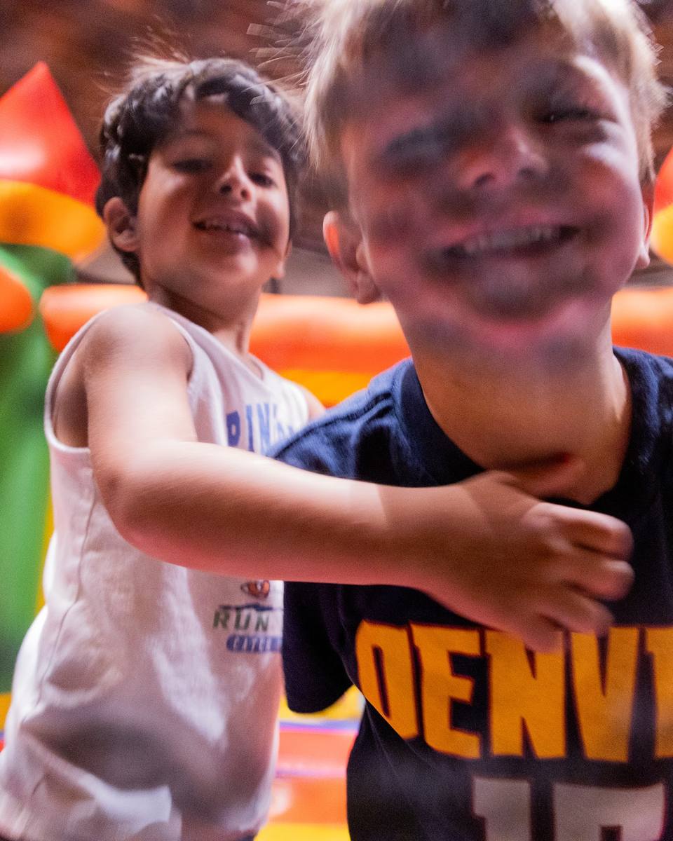 Two grinning kids horsing around in front of a bouncy castle, one in a Denver shirt
