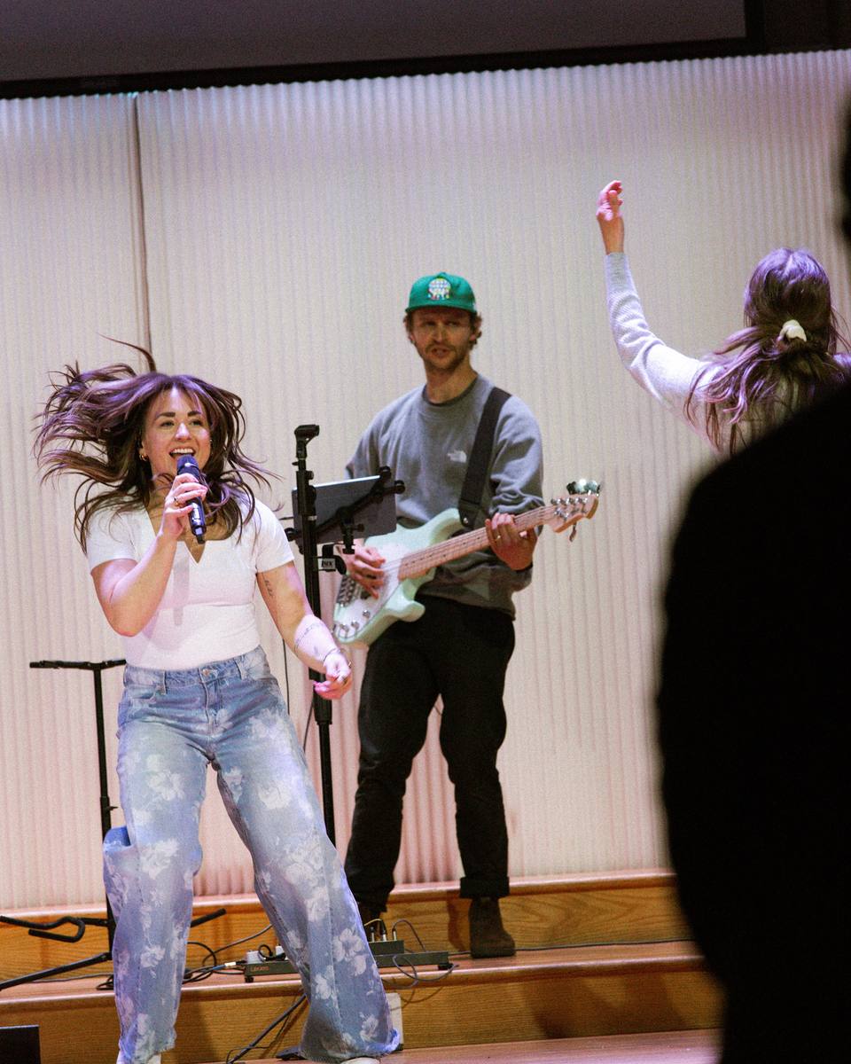 A singer with flying hair sings into a microphone on stage beside a bass player in a green cap