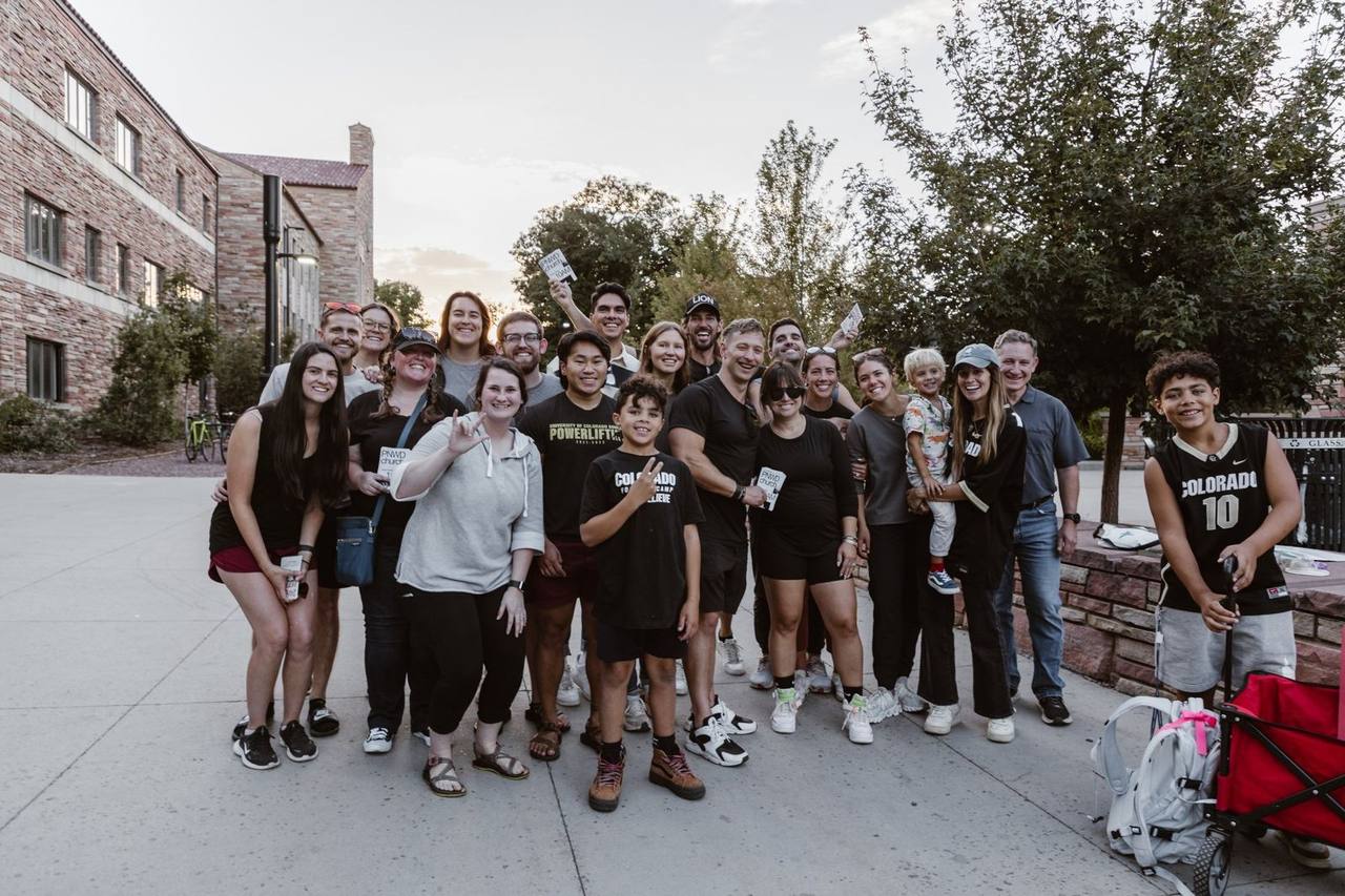 A large group posing together on a sidewalk between brick buildings at dusk, mostly young adults with a few kids and older adults among them